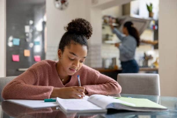 Young girl focused on completing schoolwork at home while a parent works in the background.