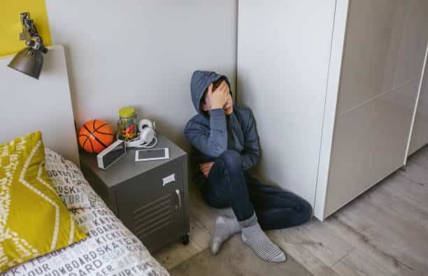 Unrecognizable depressed male teenager with his hand on his head sitting on the floor in his bedroom
